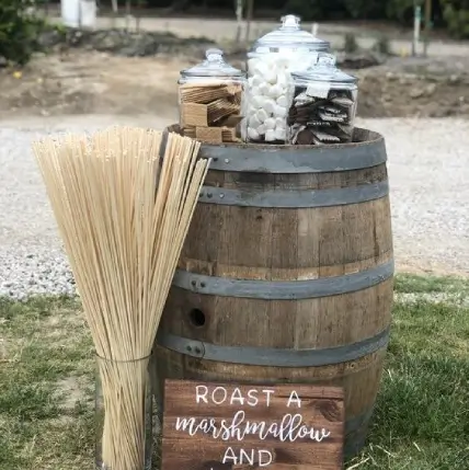 a-mini-barrel-smores-bar-with-a-sign-some-sticks-cookies-marshmallows-and-chocolate-in-jars-for-an-outdoor-wedding-space-e1693688844416